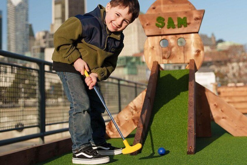 Eine Person spielt Minigolf mit einem Holz-Golfloch, auf dem 'SAN' steht, in einer Stadt. 3