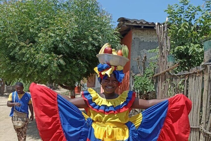 A person in a colorful costume carrying a basket on their head, with a rural setting in the background. 3