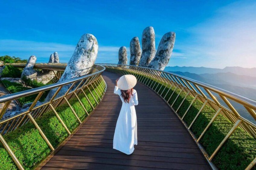 A woman in a white dress walking on a wooden boardwalk with large stone hand sculptures on either side. 4
