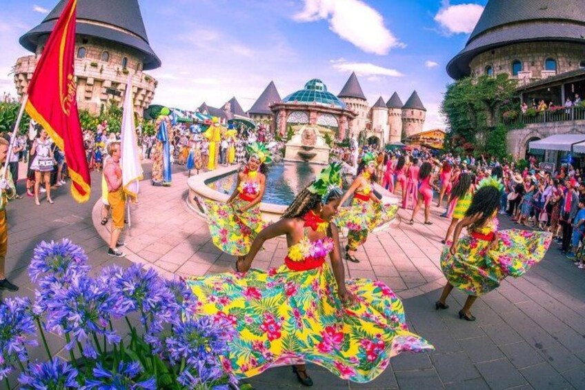 A group of dancers in colorful costumes performing in a public square with a castle-like building in the background. 2