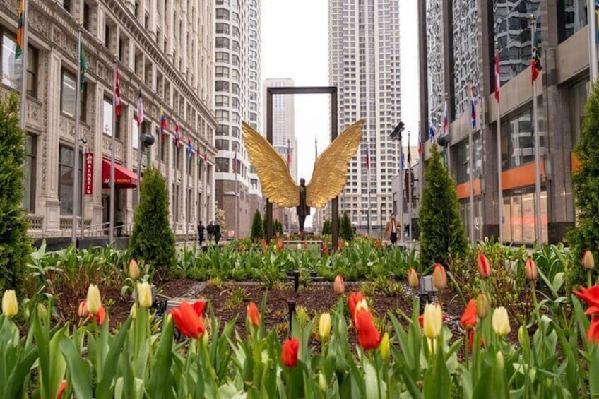 A cityscape with a large golden wing sculpture in a garden, surrounded by tulips and tall buildings. 4