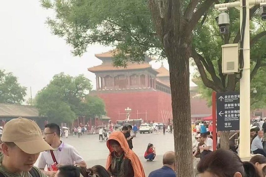Una piazza affollata con un edificio rosso che si distingue sullo sfondo, un grande albero in primo piano e segnali indicatori. 1