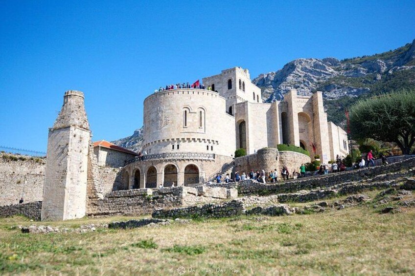A historic stone building with arches and a tower, surrounded by a grassy area and mountains. 1