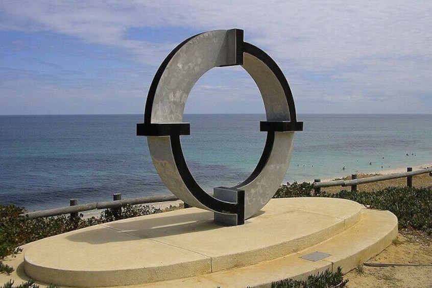 A large metal sculpture with a circular design, located on a concrete platform by the seaside. 1