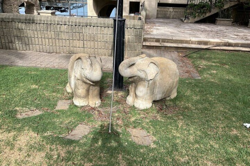 Two elephant statues on a grassy lawn with a brick wall and a staircase in the background. 4