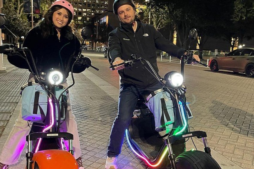 Two people on electric scooters with colorful lights, on a city street at night. 2