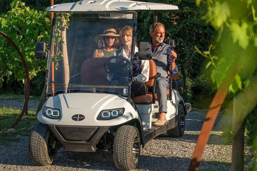 Ein Golfmobil mit zwei Personen, eine fährt und die andere hält ein Getränk, fährt auf einem Schotterweg, der von Grünflächen gesäumt wird. 1