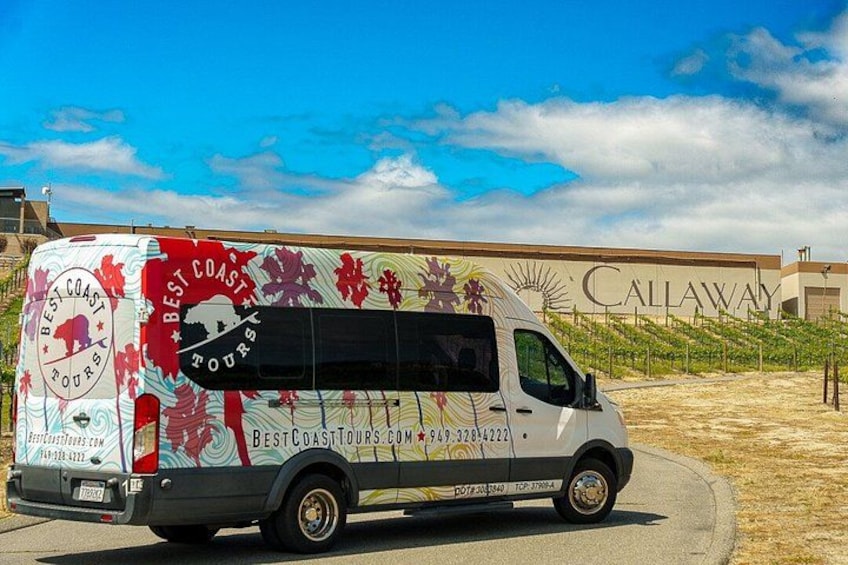 A tour bus parked in front of a building with a vineyard in the background. 2