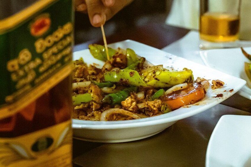 En rätt med grönsaker och kött, en flaska sås och ett glas öl på ett bord. 2
