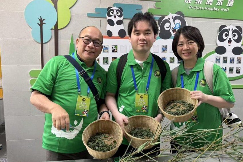 Three people in green shirts holding baskets of leaves, standing in front of a wall with panda illustrations and a sign. 1