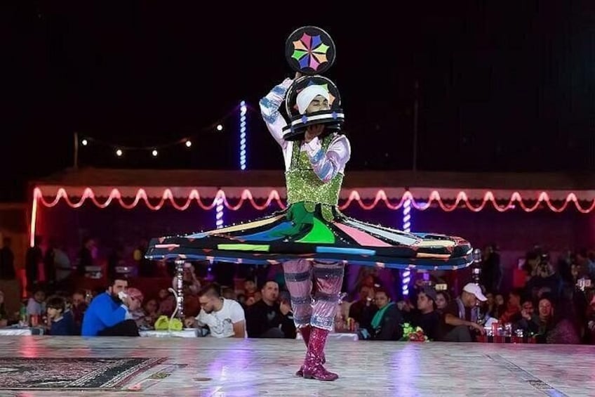 A performer in a glittery costume with a colorful headpiece, standing on a platform with a crowd in the background. 2
