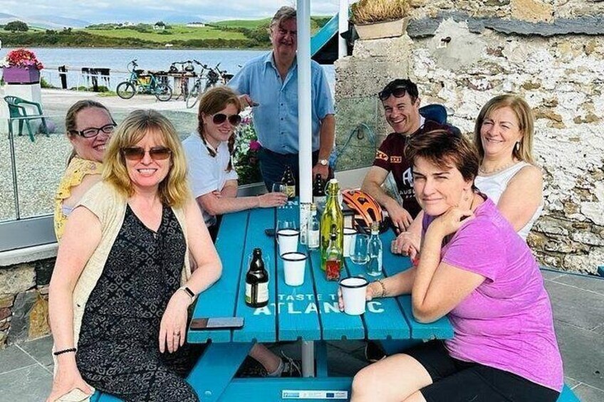 A group of people seated at a table with a blue tablecloth, outdoor setting, and a stone wall in the background. 5
