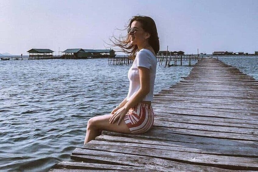 Una mujer sentada sobre un muelle de madera que se extiende sobre el agua, con bungalows sobre el agua al fondo. 1