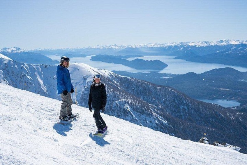 Two people snowboarding on a snowy mountain with a lake and mountains in the background. 4