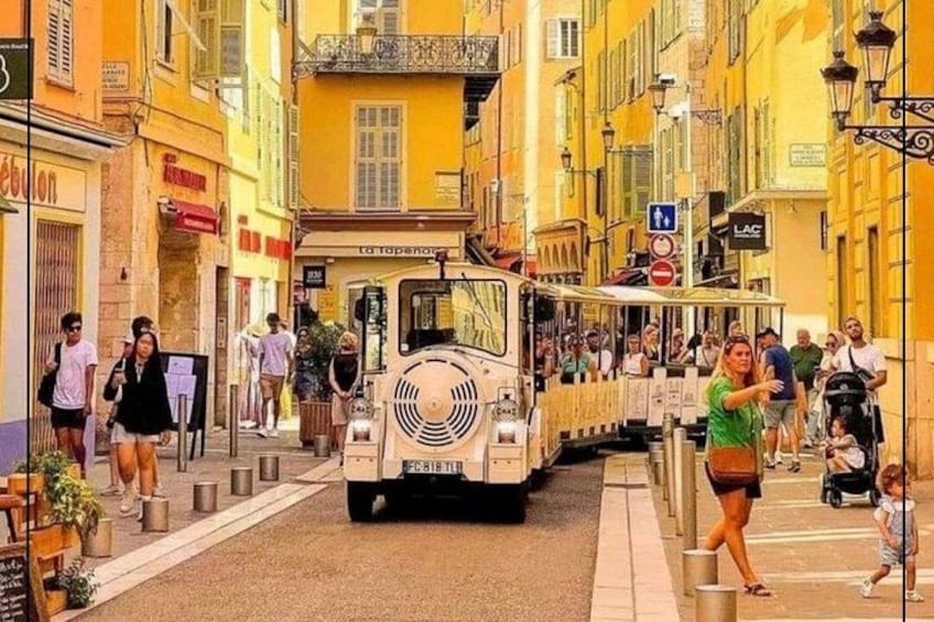 Een tram rijdt door een smalle straat met gele gebouwen, mensen lopen op de stoepen, en enkele voetgangers steken de straat over. 5