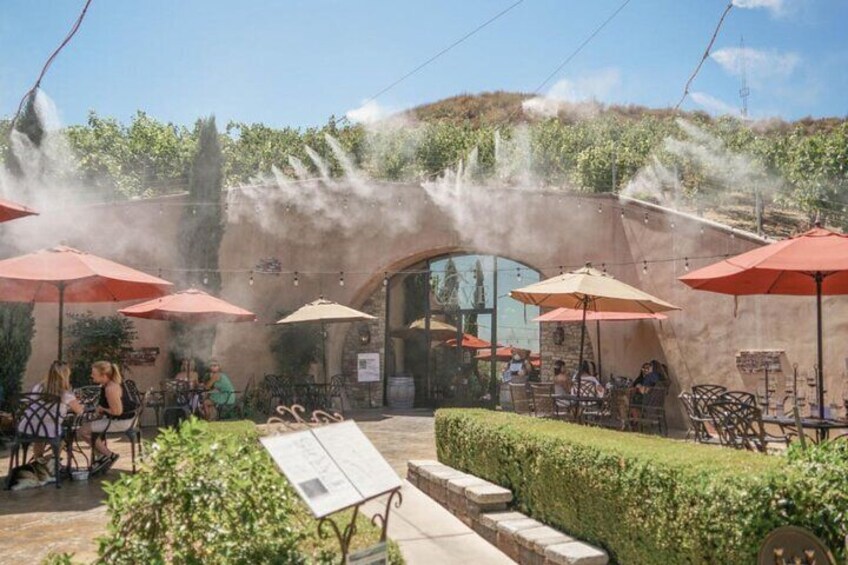 An outdoor café with tables and chairs under umbrellas, a stone archway, and a garden area. 2