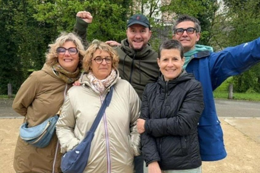 Un grupo de personas de pie al aire libre, algunas con chaquetas, con árboles y una carretera al fondo. 1