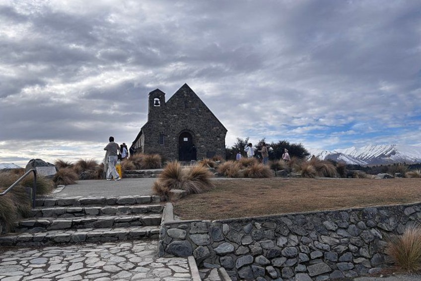 Una iglesia de piedra con un campanario, rodeada de hierba seca y arbustos, con montañas al fondo. 1
