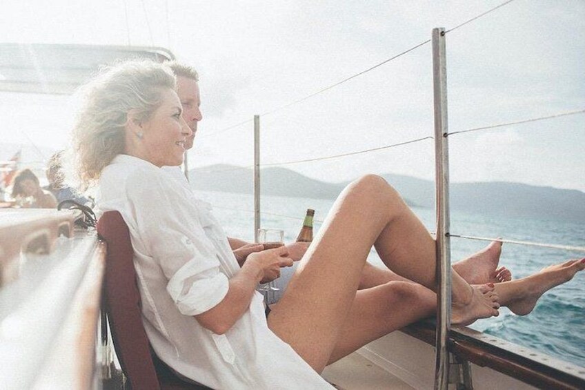 Deux personnes sont assises sur un bateau, l’une tenant une boisson. On aperçoit de l’eau et une terre lointaine en arrière-plan. 2