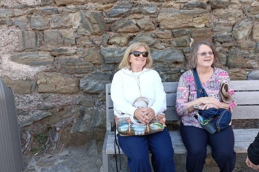 Two people sitting on a wooden bench against a stone wall. 4