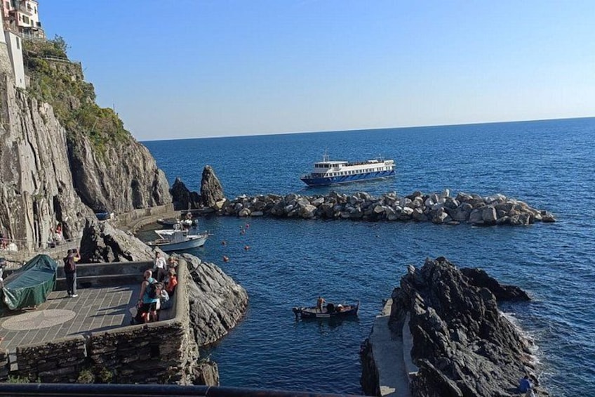 A rocky coastline with a boat docked near the shore, a pier with people, and a clear blue sky. 3