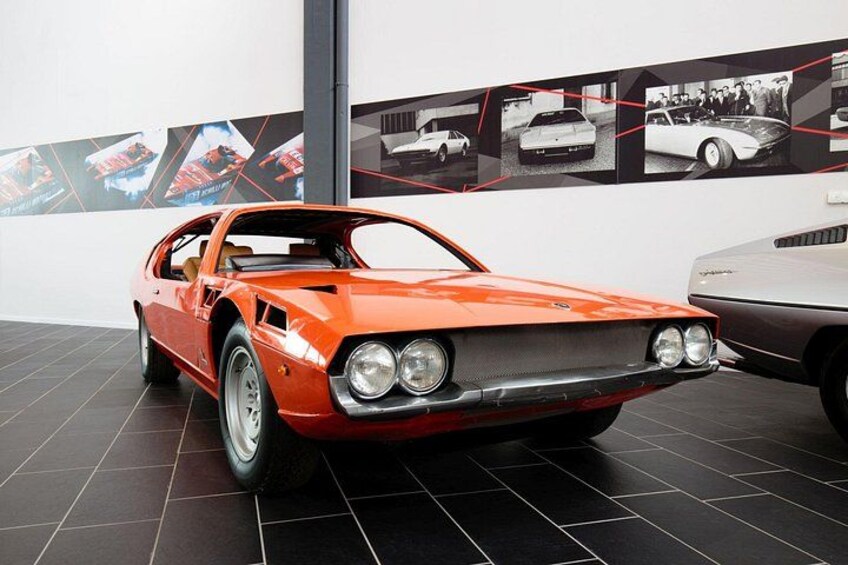 Une voiture de sport classique exposée dans un showroom, avec un sol carrelé de carreaux noirs et un mur recouvert d’affiches de voitures anciennes. 2