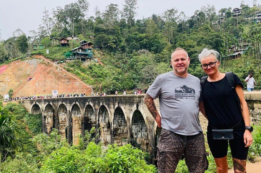 A man and woman standing in front of a bridge with multiple arches, surrounded by lush greenery and hills. 1