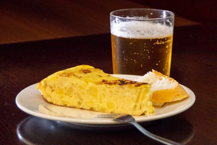 A slice of quiche on a plate with a fork, and a glass of beer in the background. 4