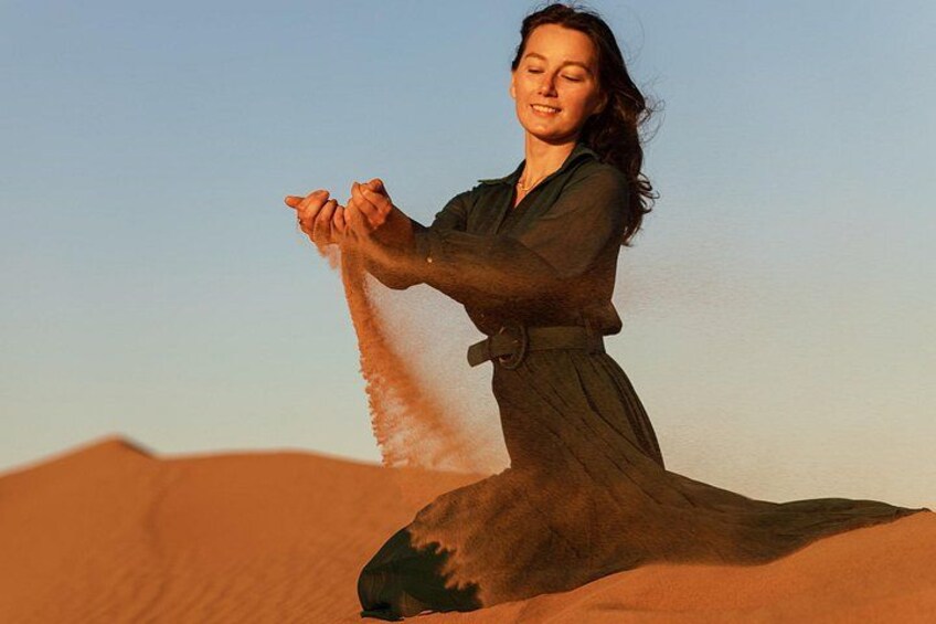 A person in a flowing dress kneeling on a sand dune, holding sand in their hands. 1