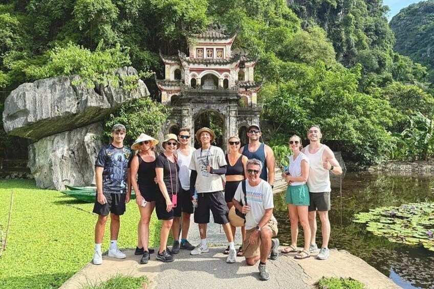 Un groupe de personnes posant devant un pavillon chinois traditionnel, au bord d’un étang bordé d’une végétation luxuriante, avec un grand rocher en arrière-plan. 1