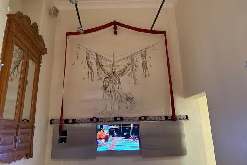 A room with a large tapestry depicting a figure with a horse, a wooden cabinet, and a television screen. 2