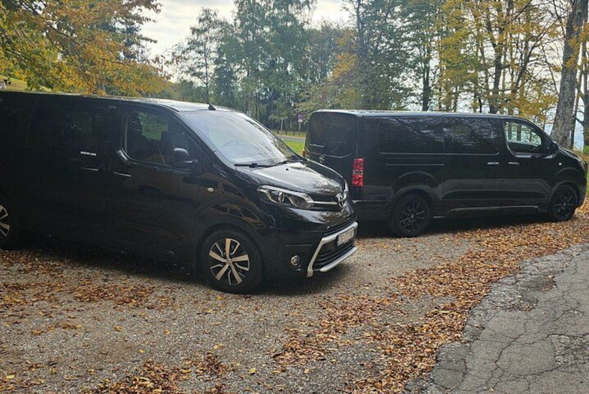 Twee zwarte voertuigen geparkeerd op een grindpad, omgeven door bomen met herfstbladeren en vallend blad. 2