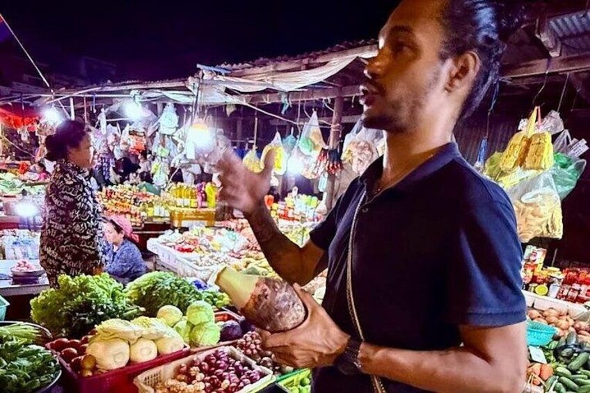 A person standing at a market stall with various vegetables and fruits. 2