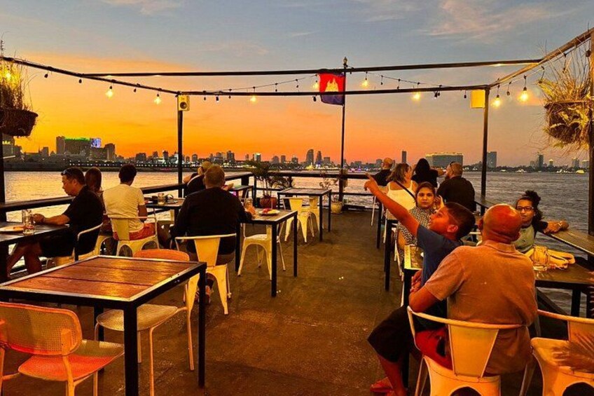 A waterfront dining area with tables and chairs, overlooking a river with a city skyline in the background. 5