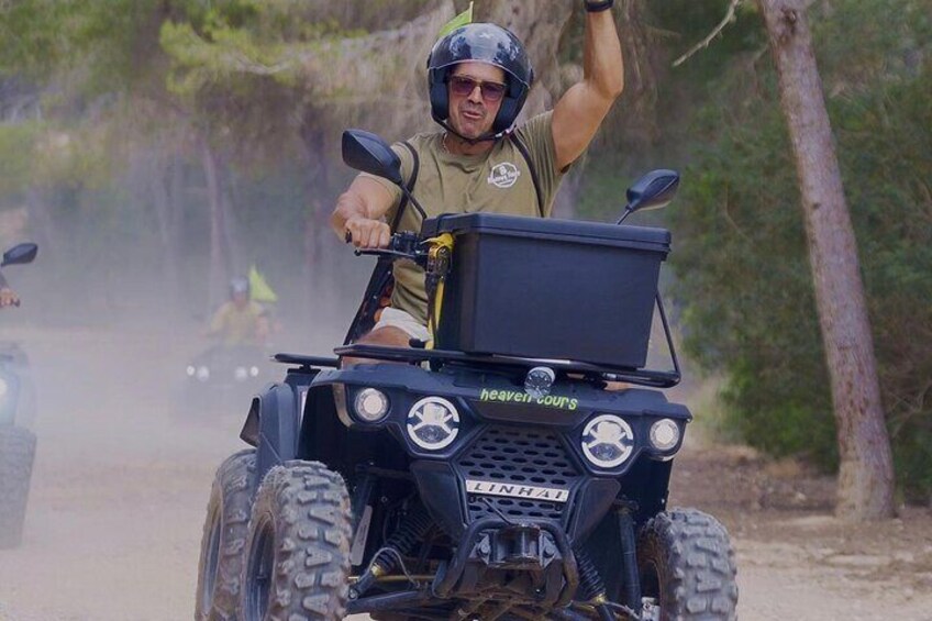 Une personne conduit un quad noir sur un chemin de terre bordé d’arbres. Le quad est équipé d’un coffre de rangement sur lequel on peut lire l’inscription « Heaven Tours ». 1