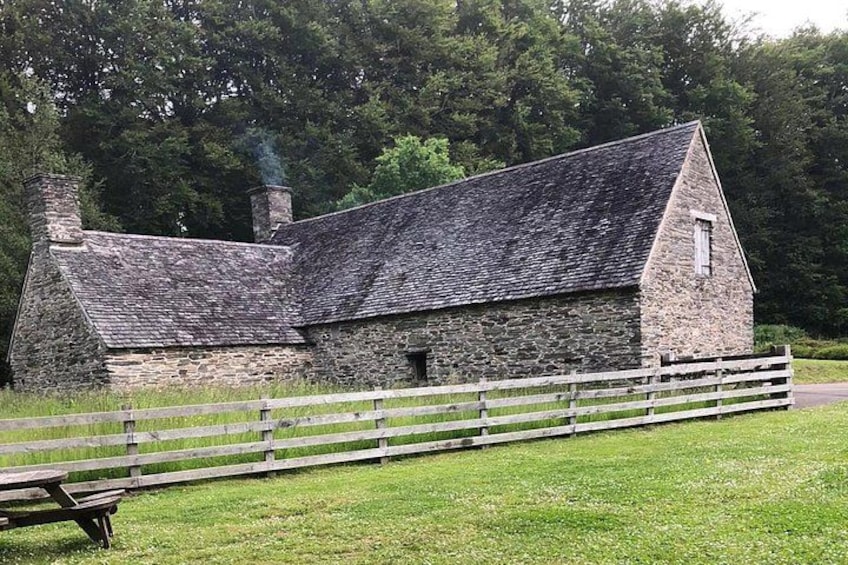 Ein Steinhaus mit geneigtem Dach, umgeben von einem Holzzaun, in einer grünen Umgebung mit Bäumen im Hintergrund. 2
