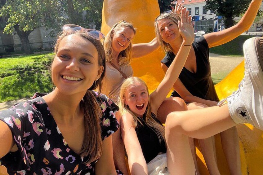 A group of people sitting on a yellow structure in a park with greenery and buildings in the background. 2