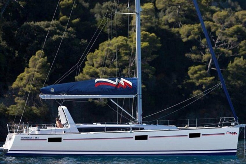 Un velero blanco con toldo azul y logotipo rojo y blanco navegando sobre aguas tranquilas, con un bosque al fondo. 5