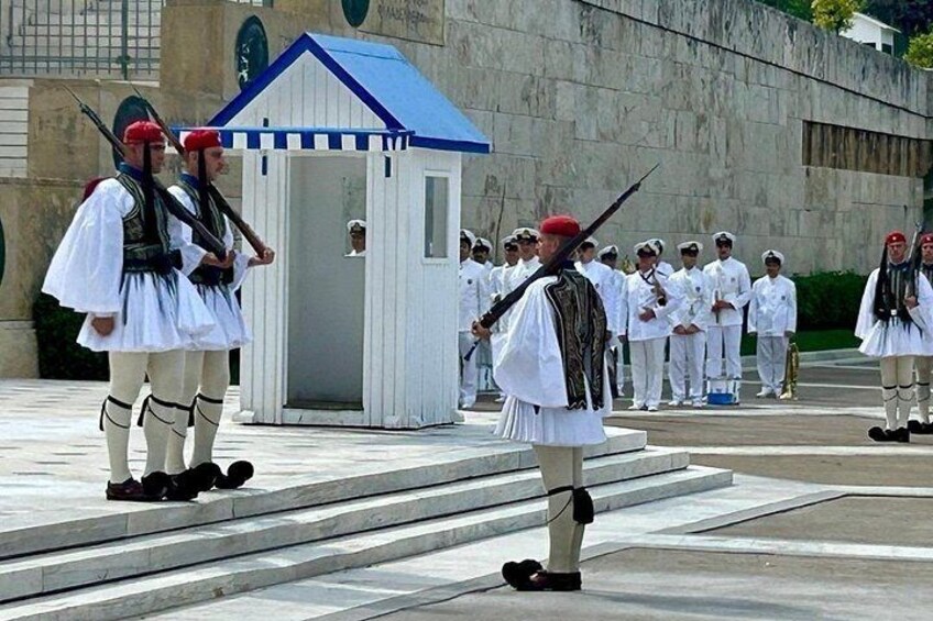 A ceremonial guard in traditional attire, a small building, and a group of uniformed personnel. 1