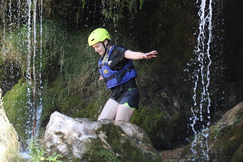 Une personne portant un casque et un gilet de sauvetage, debout sur un rocher près d’une cascade. 2