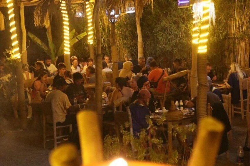 Area pranzo all'aperto con persone sedute a tavoli, circondata da palme e illuminata da luci decorative. 4