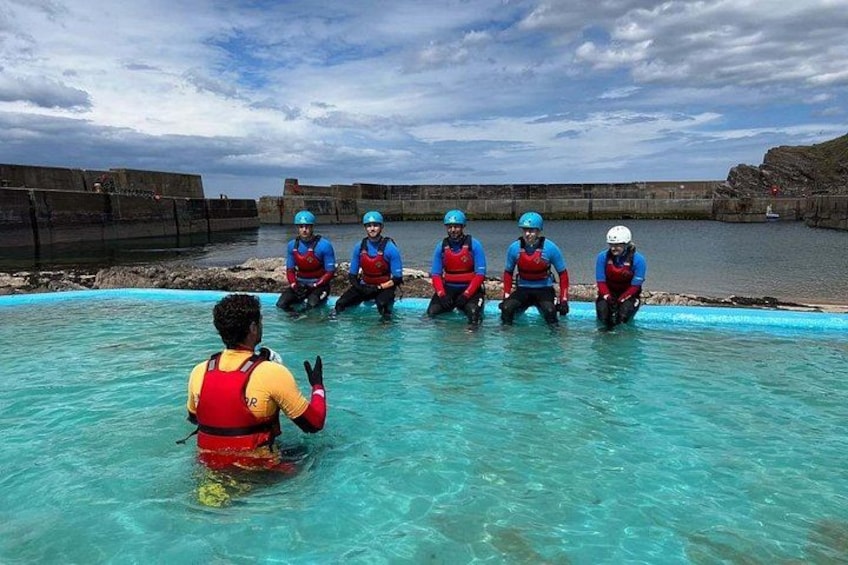 A group of people in a pool wearing life jackets and helmets, with a rocky shoreline in the background. 5