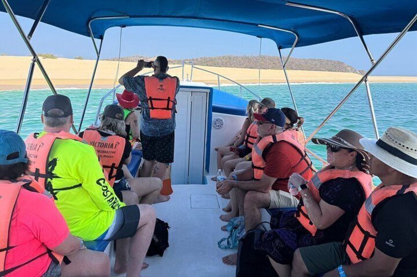 A group of people on a boat wearing life jackets, with a sandy beach and clear blue water in the background. 2