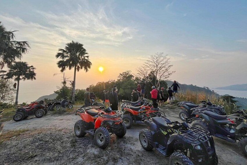 Solnedgang med ATV'er parkeret på en jordvej, palmer og et bakket landskab. 3