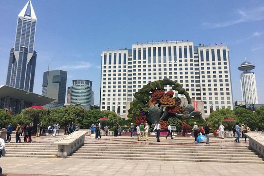 Una piazza pubblica con un’ampia composizione floreale, edifici moderni e una torre sullo sfondo. 1