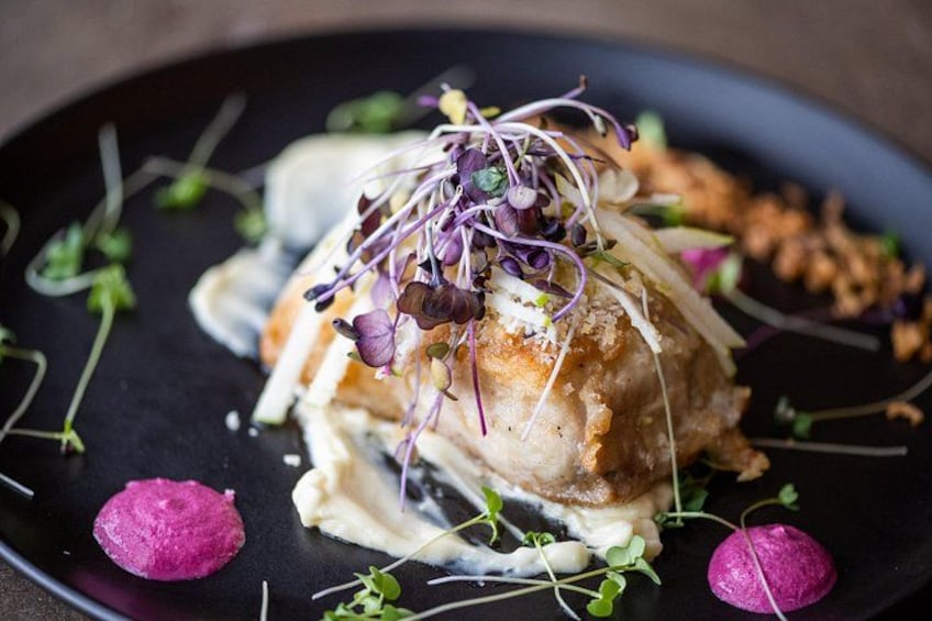 A dish with a piece of meat, possibly chicken, topped with sprouts and a creamy sauce, served on a black plate with some greens and purple elements. 3