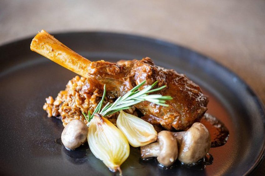 A dish with a lamb shank, mushrooms, and a sprig of rosemary on a black plate. 4
