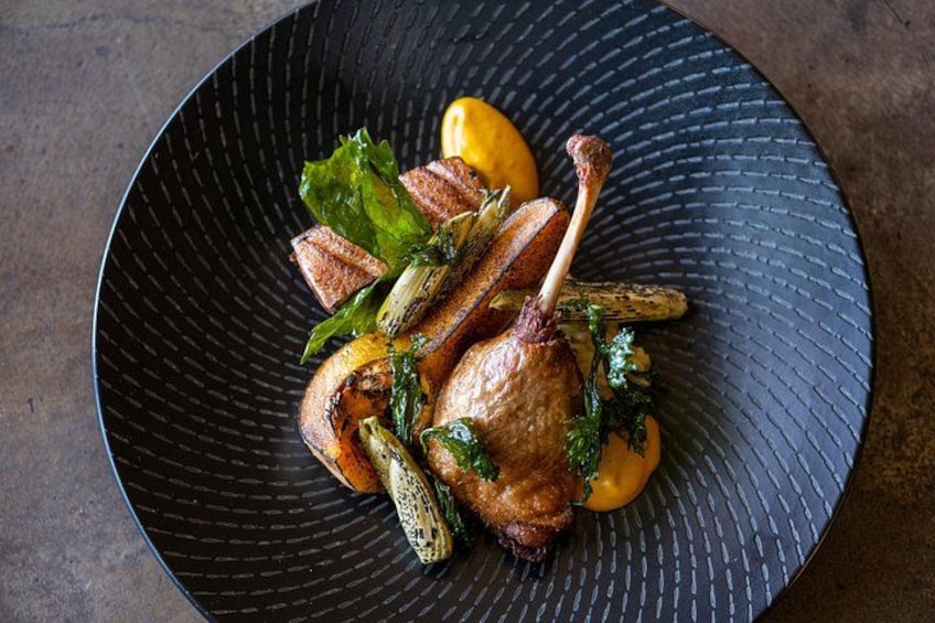 A roasted duck leg with vegetables on a black plate. 2