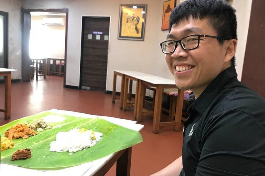 A person sitting at a table with a meal on a banana leaf, a wooden table, and a room with chairs and a door in the background. 2