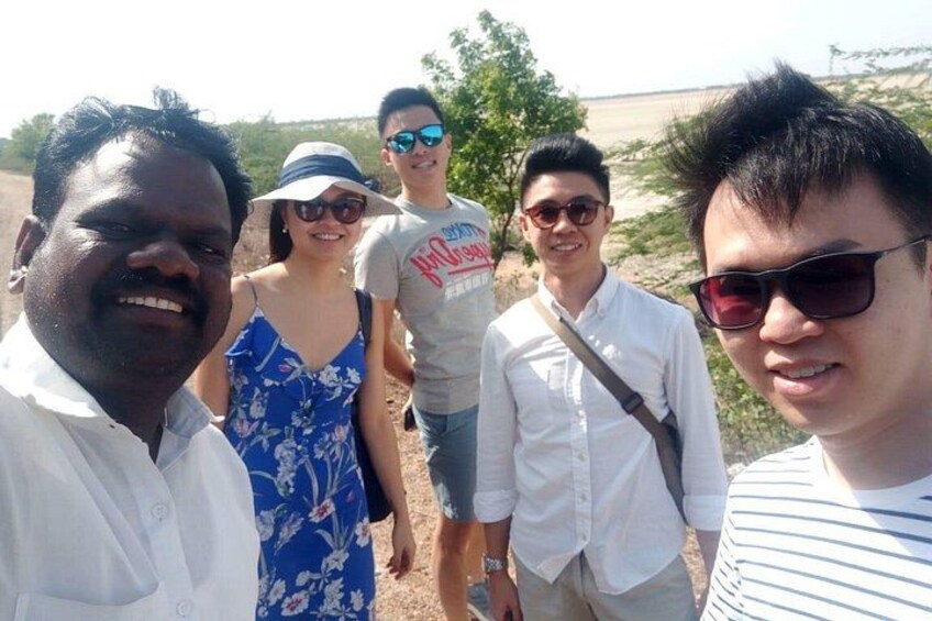 A group of people standing outdoors, possibly on a beach or near a body of water, with a clear sky and sparse vegetation in the background. 5